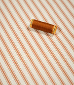 JF Ticking Burnt Orange fabric styled with a coordinating burnt orange thread spool, highlighting the striped design and textured cream weave.