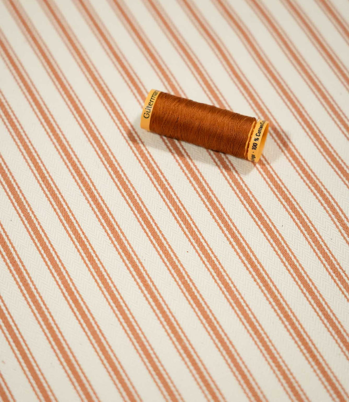 JF Ticking Burnt Orange fabric styled with a coordinating burnt orange thread spool, highlighting the striped design and textured cream weave.