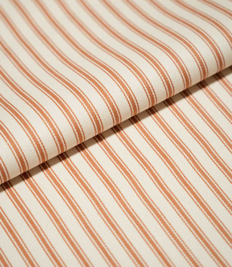 Close-up of folded JF Ticking Burnt Orange fabric showing the repeating burnt orange stripe pattern and subtle herringbone woven texture.