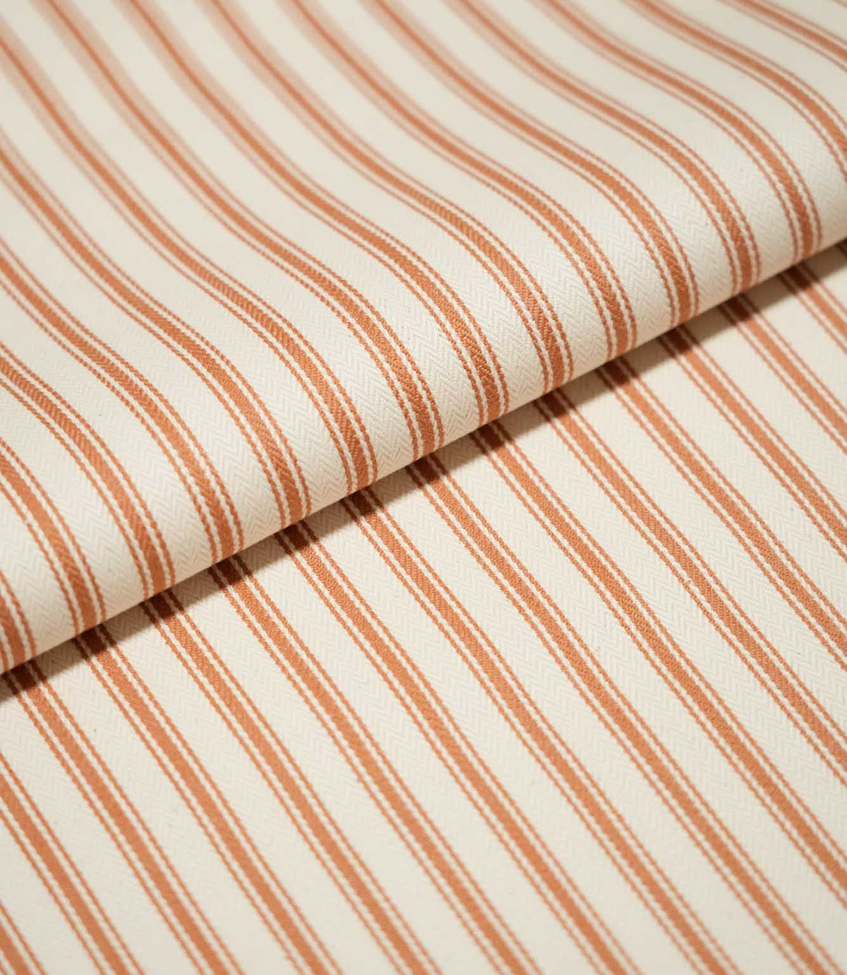 Close-up of folded JF Ticking Burnt Orange fabric showing the repeating burnt orange stripe pattern and subtle herringbone woven texture.
