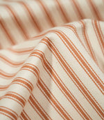Detailed close-up of JF Ticking Burnt Orange fabric showing the woven burnt orange ticking stripes, warm cream ground and fine herringbone texture.