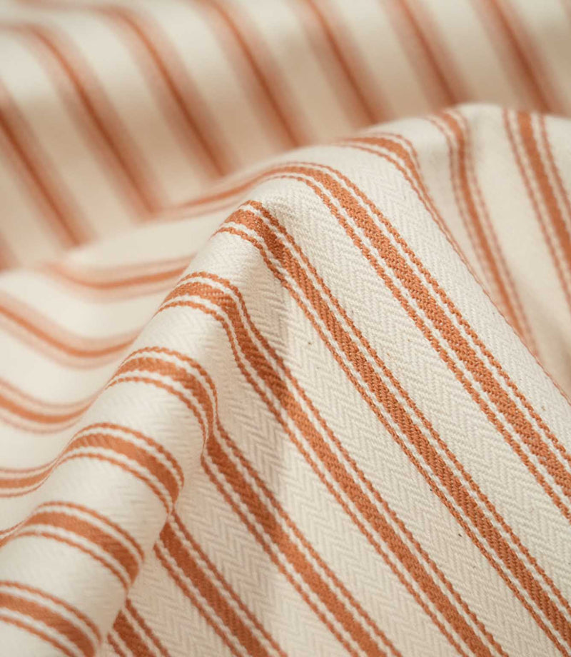 Detailed close-up of JF Ticking Burnt Orange fabric showing the woven burnt orange ticking stripes, warm cream ground and fine herringbone texture.