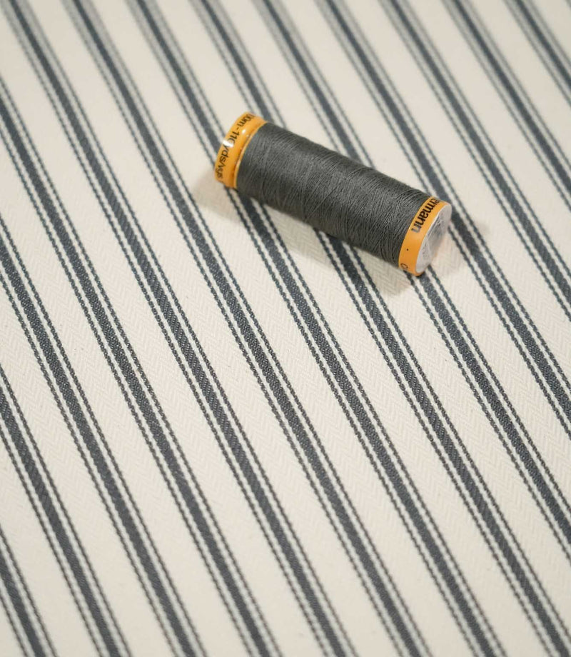 JF Ticking Grey fabric styled with a coordinating grey thread spool, highlighting the classic stripe design and subtle herringbone weave.