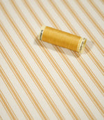 JF Ticking Ochre fabric styled with a coordinating ochre thread spool, highlighting the classic striped pattern and textured woven surface.