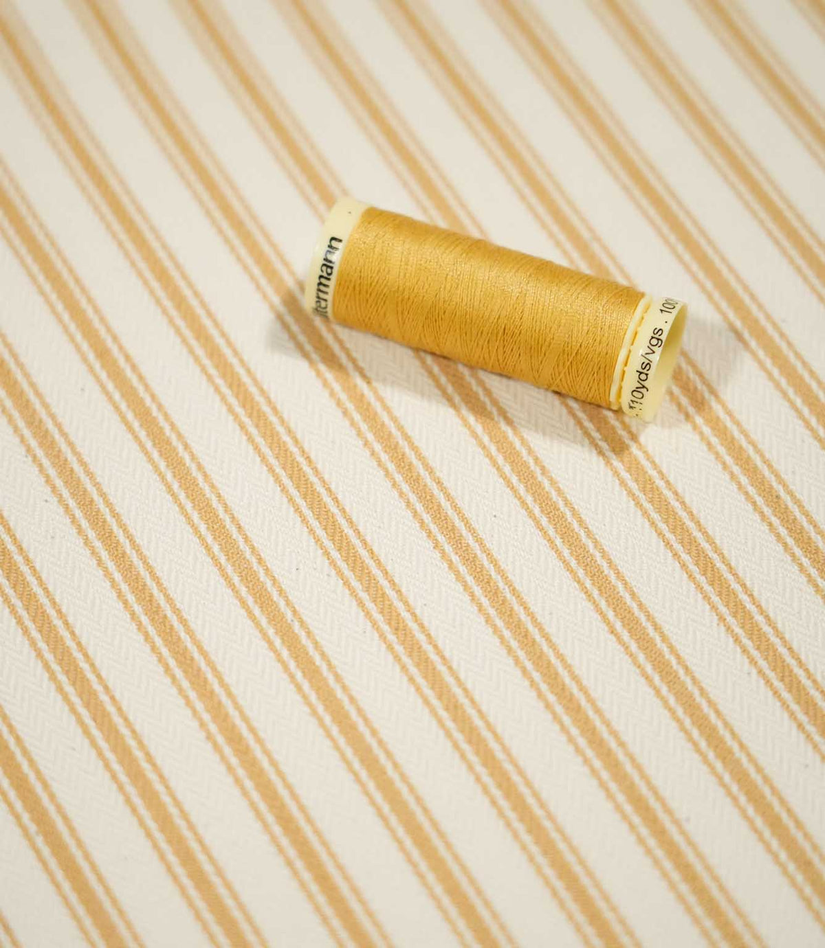JF Ticking Ochre fabric styled with a coordinating ochre thread spool, highlighting the classic striped pattern and textured woven surface.