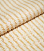 Close-up of folded JF Ticking Ochre fabric showing warm ochre ticking stripes against a cream ground with a subtle herringbone weave.