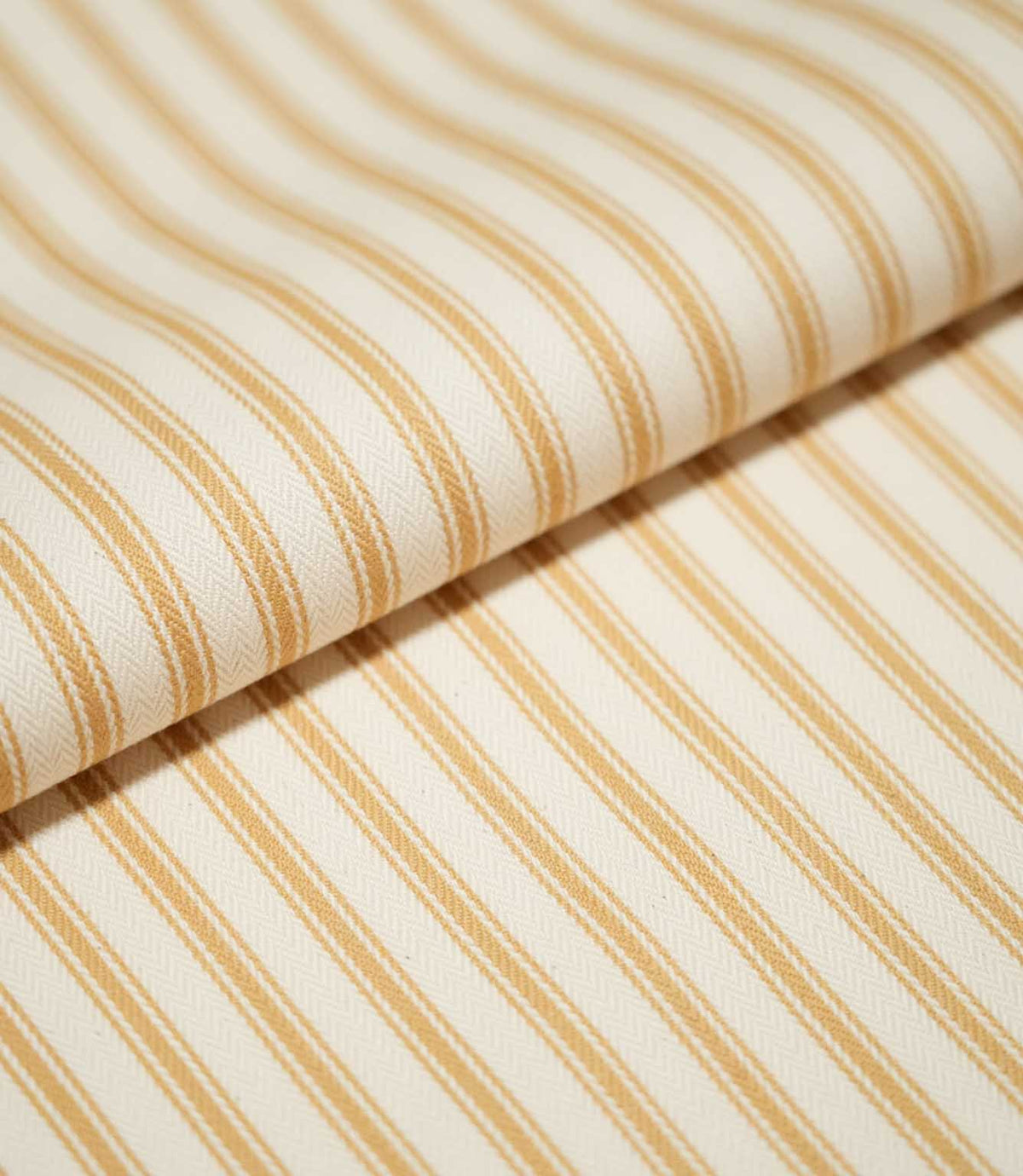Close-up of folded JF Ticking Ochre fabric showing warm ochre ticking stripes against a cream ground with a subtle herringbone weave.