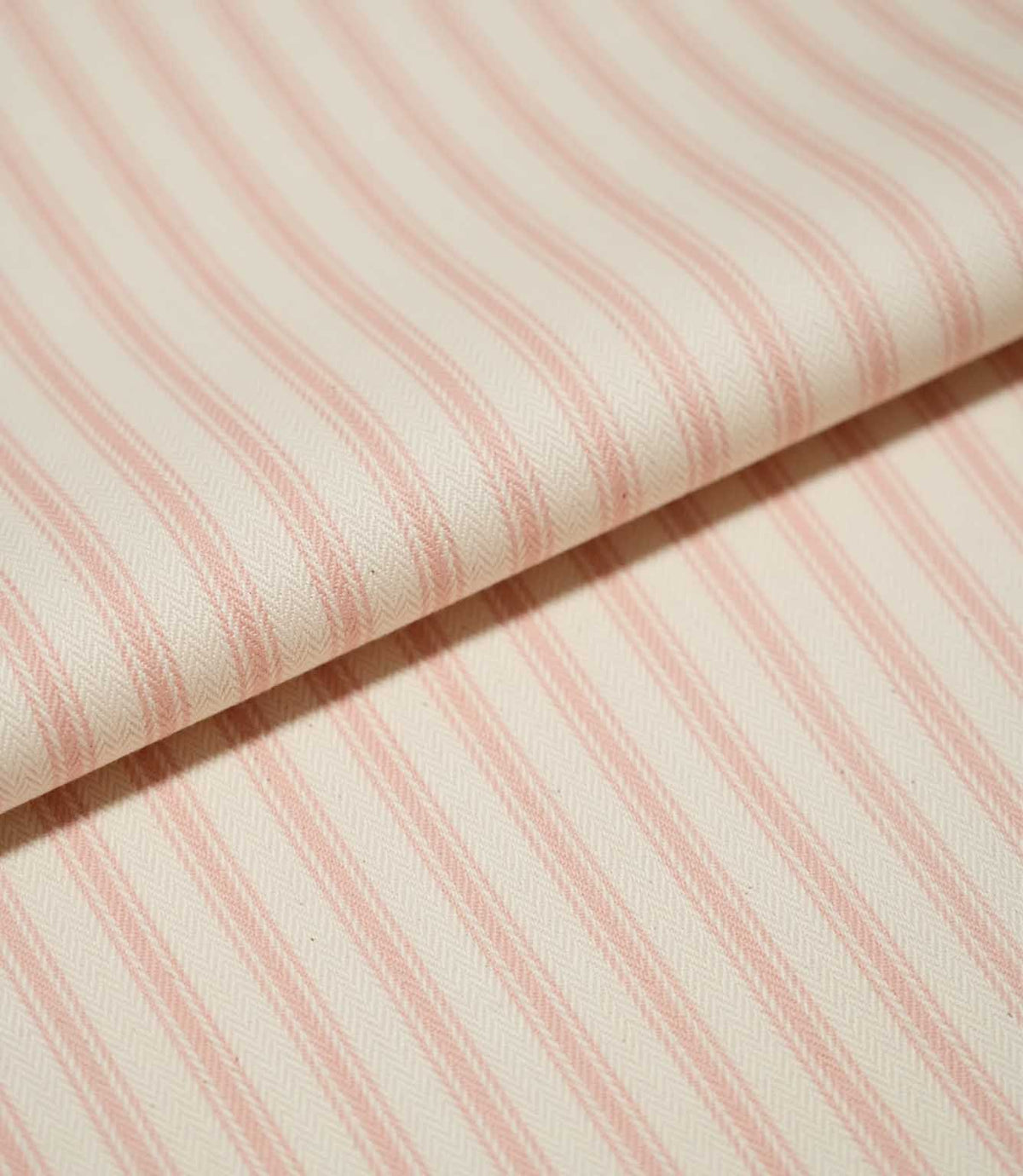 Close-up of folded JF Ticking Pink fabric showing soft pink ticking stripes and the fine herringbone texture of the cream woven ground.