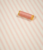 JF Ticking Pink fabric displayed with a coordinating pink thread spool, highlighting the repeating pink stripes and subtle herringbone weave.