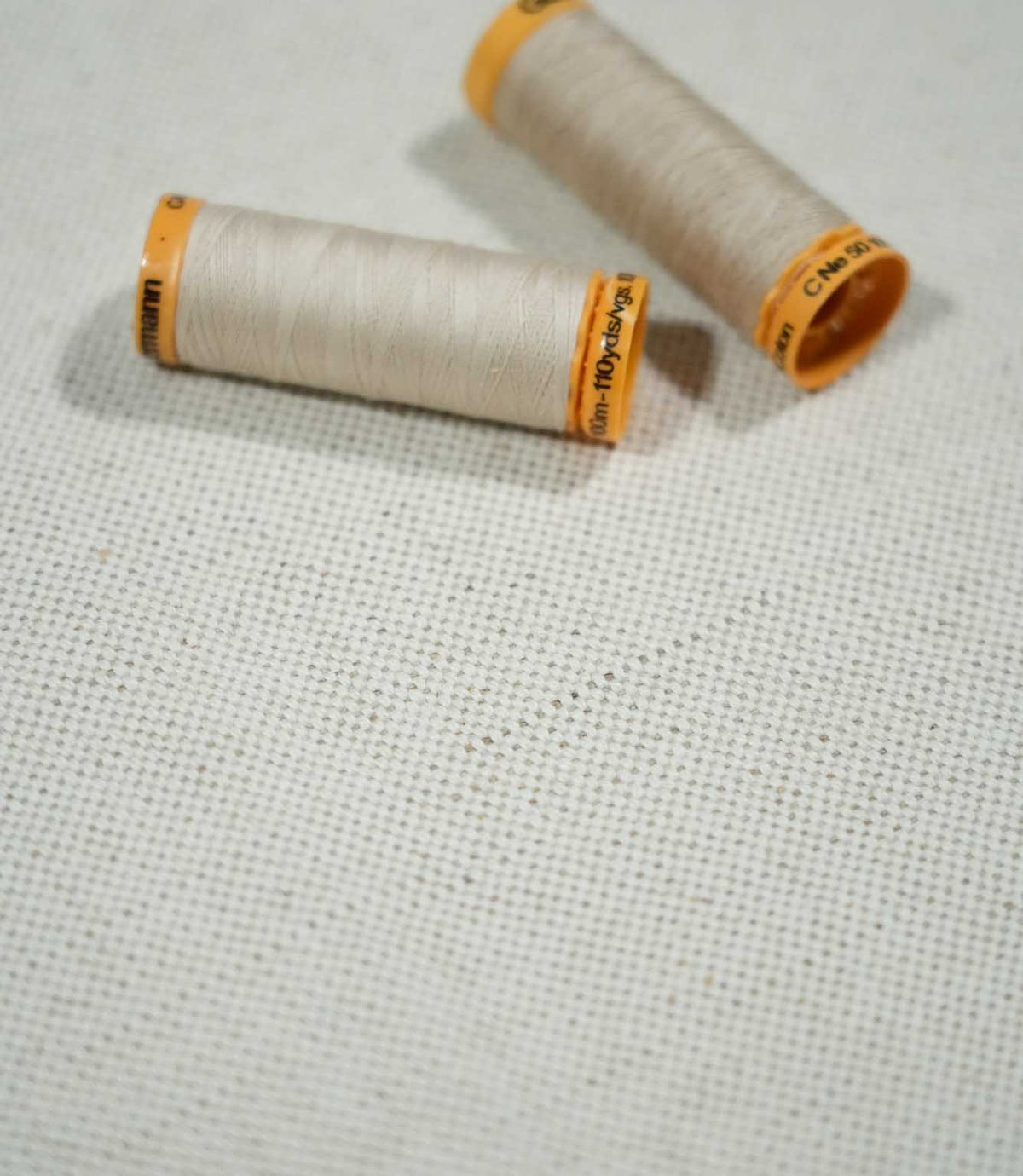 JF Ticking Plain Natural fabric photographed with two cream sewing thread spools for scale, highlighting the textured weave and subtle natural beige fibres.