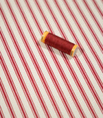 JF Ticking Red fabric styled with a coordinating red thread spool, highlighting the classic red ticking stripes and subtle cream herringbone weave.