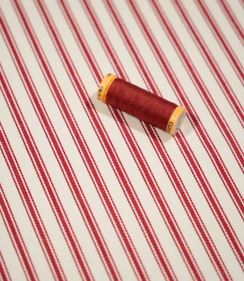 JF Ticking Red fabric styled with a coordinating red thread spool, highlighting the classic red ticking stripes and subtle cream herringbone weave.