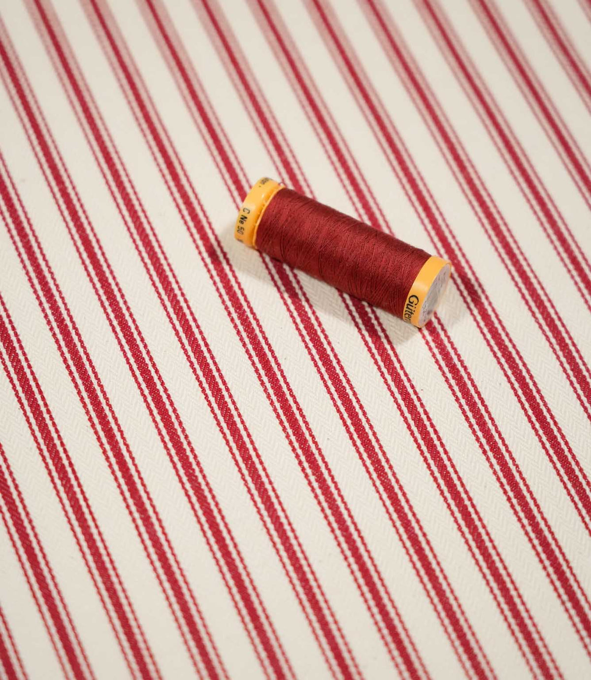 JF Ticking Red fabric styled with a coordinating red thread spool, highlighting the classic red ticking stripes and subtle cream herringbone weave.