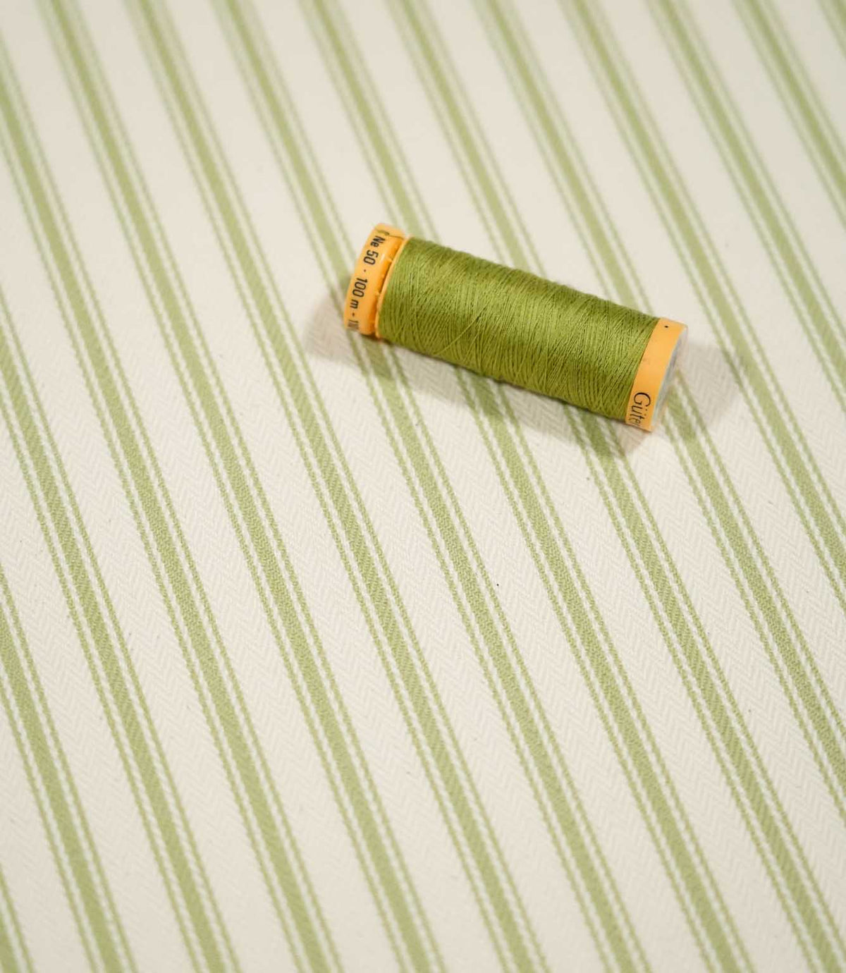JF Ticking Sage Green fabric styled with a coordinating green thread spool, highlighting the striped design and subtle cream herringbone weave.