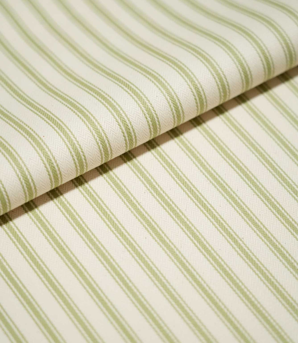 Close-up of folded JF Ticking Sage Green fabric showing the repeating sage green ticking stripes and fine herringbone texture of the cream background.