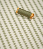 JF Ticking Sap Green fabric styled with a coordinating olive green thread spool, highlighting the classic striped design and subtle herringbone weave.