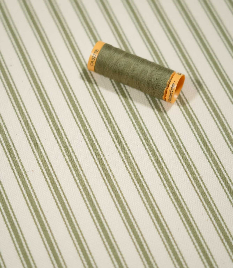JF Ticking Sap Green fabric styled with a coordinating olive green thread spool, highlighting the classic striped design and subtle herringbone weave.