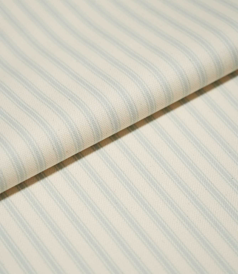 Close-up of folded JF Ticking Seaspray fabric showing soft blue ticking stripes and the detailed herringbone weave of the cream background.