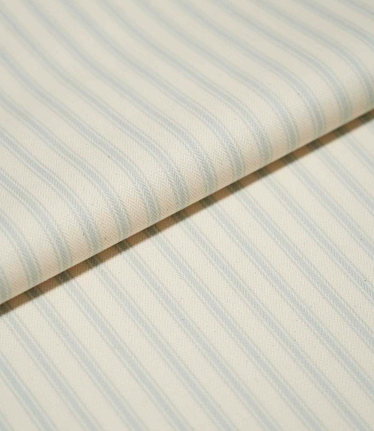 Close-up of folded JF Ticking Seaspray fabric showing soft blue ticking stripes and the detailed herringbone weave of the cream background.