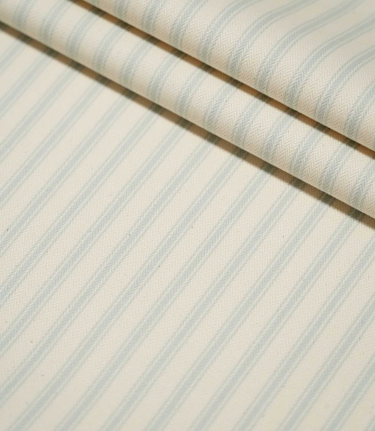 JF Ticking Seaspray striped fabric arranged in neat folds, highlighting the repeating pale blue stripes and subtle textured herringbone weave.