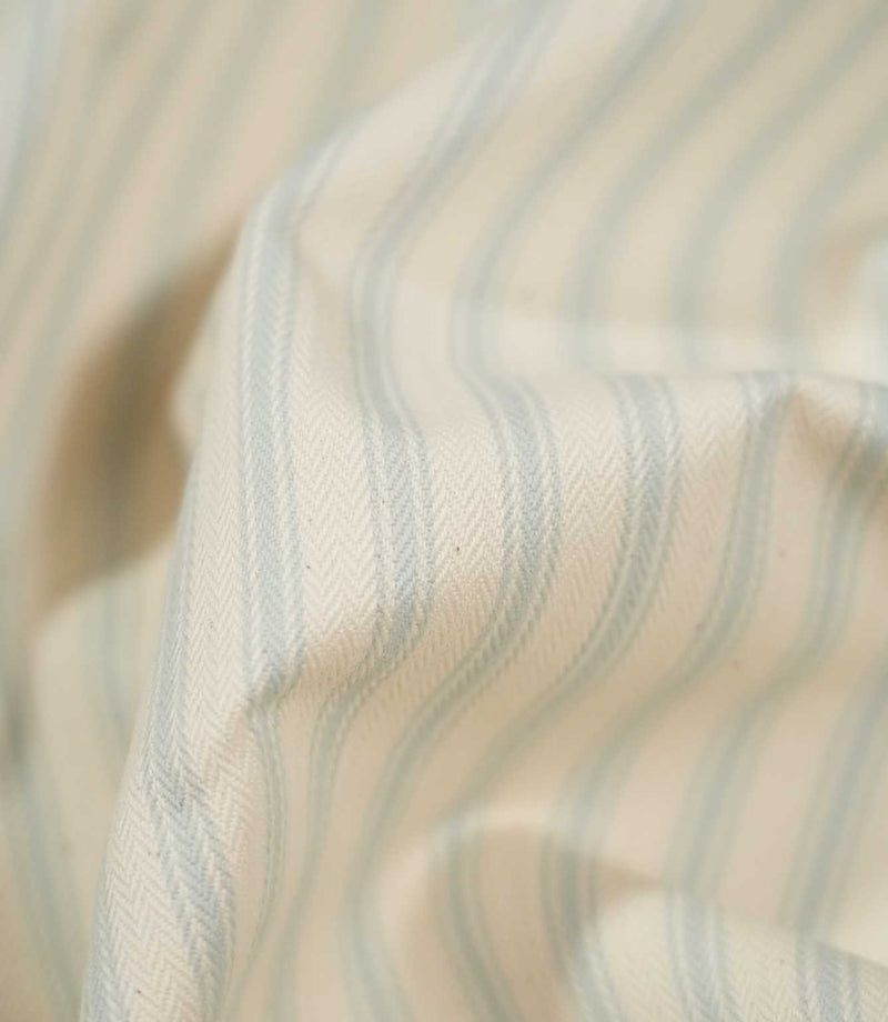 Detailed close-up of JF Ticking Seaspray fabric draped over an edge, highlighting the pale blue stripes, cream ground and fine herringbone woven texture.