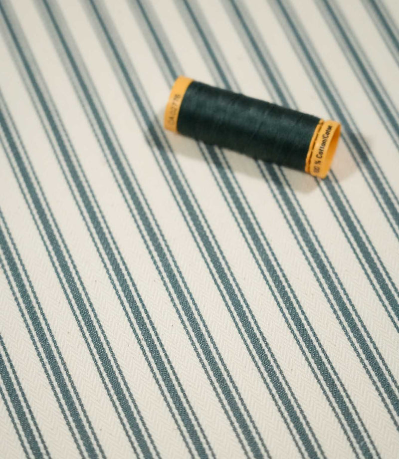 JF Ticking Teal fabric styled with a coordinating dark teal thread spool, highlighting the classic striped design and textured woven surface.