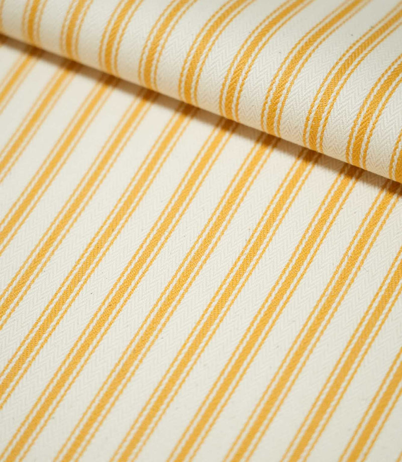 Close-up of JF Ticking Turmeric fabric folded over itself, highlighting the golden yellow ticking stripes, warm cream background and subtle herringbone woven texture.