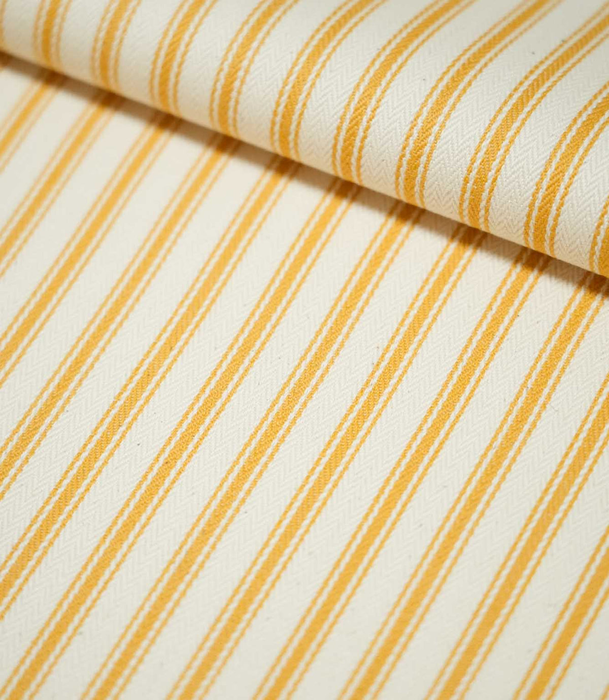 Close-up of JF Ticking Turmeric fabric folded over itself, highlighting the golden yellow ticking stripes, warm cream background and subtle herringbone woven texture.