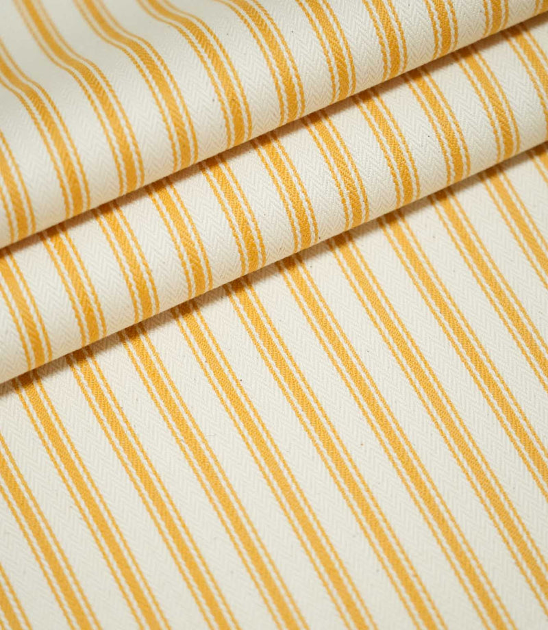 JF Ticking Turmeric fabric arranged in overlapping folds, showing the repeating golden yellow ticking stripes, fine parallel lines and textured cream herringbone weave.