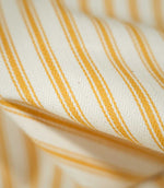 Macro photograph of JF Ticking Turmeric fabric showing the fine woven texture, natural cream base and golden yellow ticking stripes, with wider central lines bordered by narrower stripes.