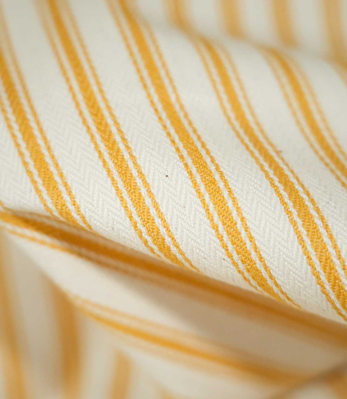 Macro photograph of JF Ticking Turmeric fabric showing the fine woven texture, natural cream base and golden yellow ticking stripes, with wider central lines bordered by narrower stripes.