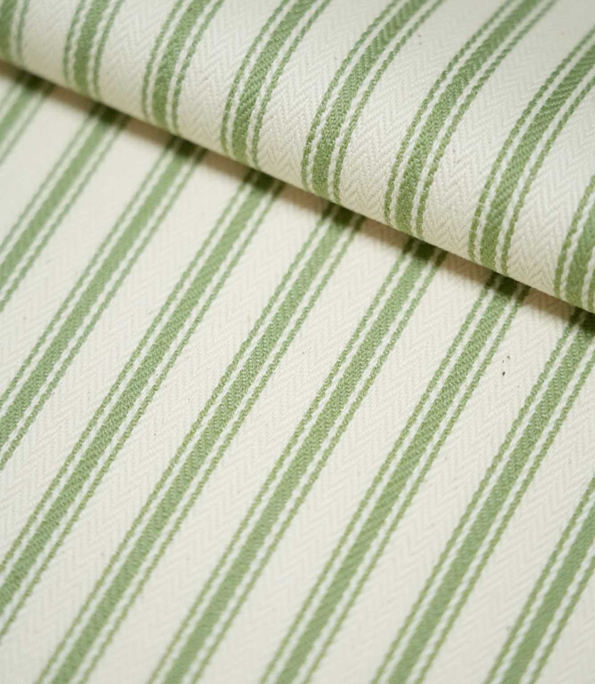 Close-up of JF Ticking Warm Green fabric folded over itself, highlighting the sage green ticking stripes, natural cream background and subtle herringbone woven texture.