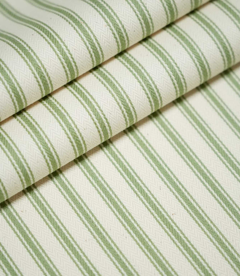 JF Ticking Warm Green fabric arranged in overlapping folds, showing the repeating sage green vertical stripes, fine contrasting lines and textured cream herringbone weave.