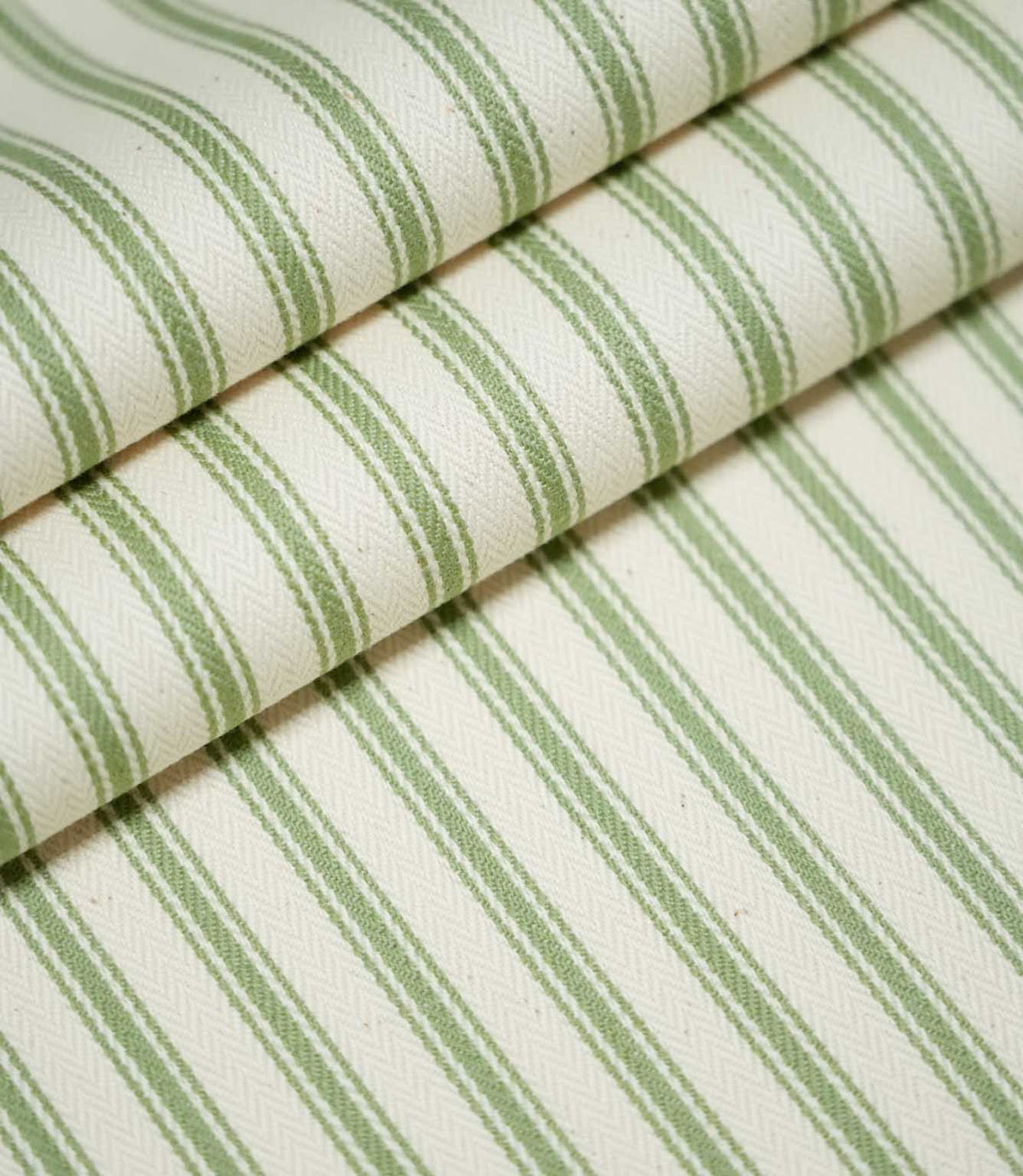 JF Ticking Warm Green fabric arranged in overlapping folds, showing the repeating sage green vertical stripes, fine contrasting lines and textured cream herringbone weave.