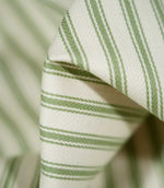Macro photograph of JF Ticking Warm Green fabric showing the fine herringbone weave, soft cream base and sage green ticking stripes, with wider central lines bordered by narrower stripes.