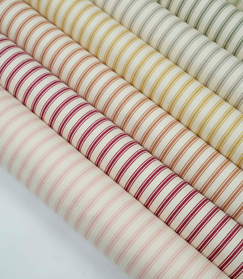 Collection of ticking stripe fabrics in blush pink, red, burnt orange, mustard, grey and sage green colourways with woven herringbone texture.