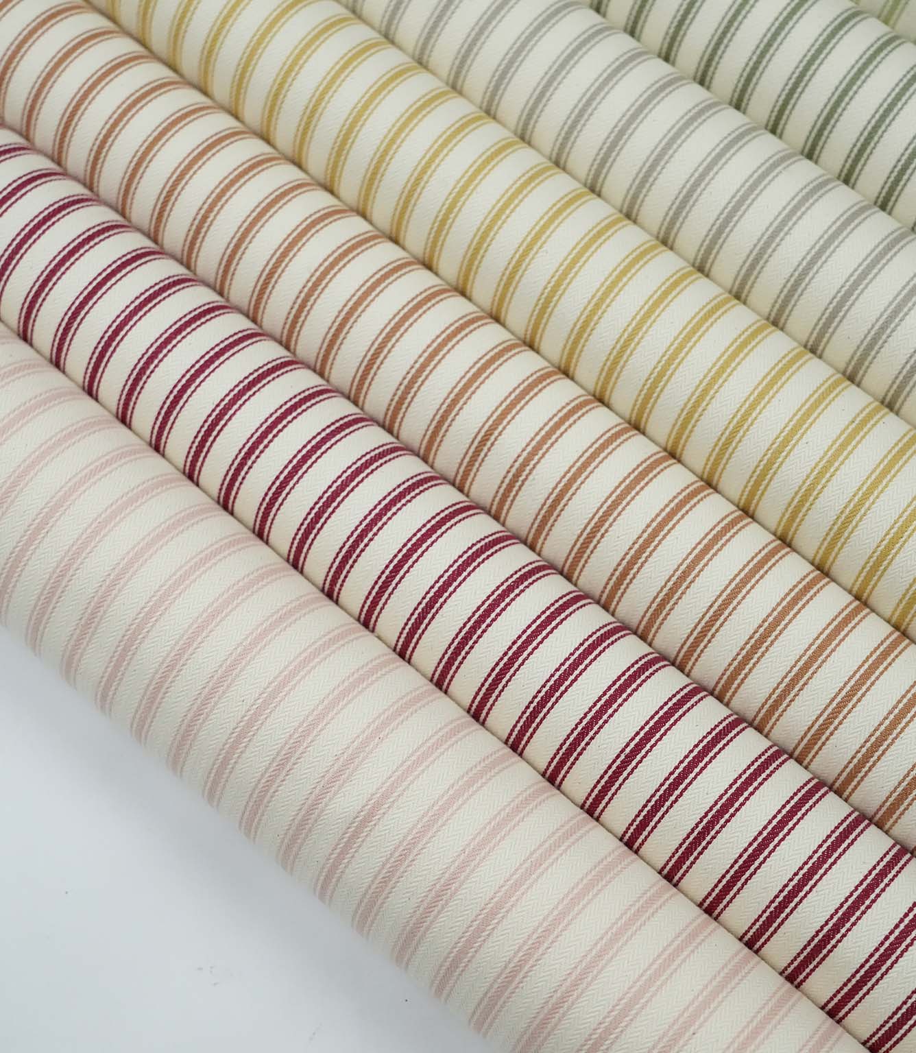 Collection of ticking stripe fabrics in blush pink, red, burnt orange, mustard, grey and sage green colourways with woven herringbone texture.