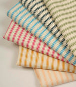 Stacked selection of ticking stripe fabrics in olive green, indigo blue, sky blue, pink, mustard yellow, and peach tones on ivory herringbone woven cotton fabric.