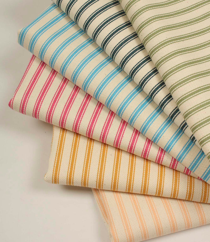 Stacked selection of ticking stripe fabrics in olive green, indigo blue, sky blue, pink, mustard yellow, and peach tones on ivory herringbone woven cotton fabric.
