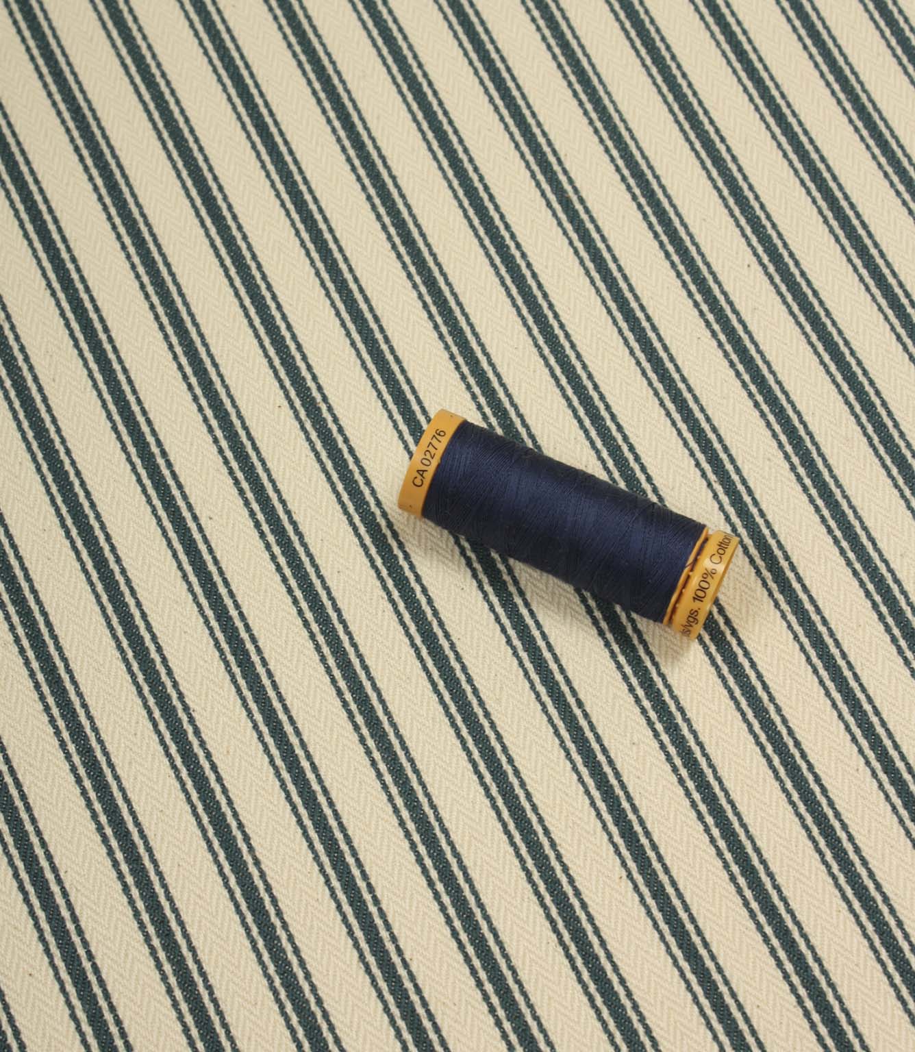 Indigo ticking stripe fabric displayed flat with a coordinating navy thread spool, showcasing the traditional striped design and woven texture.