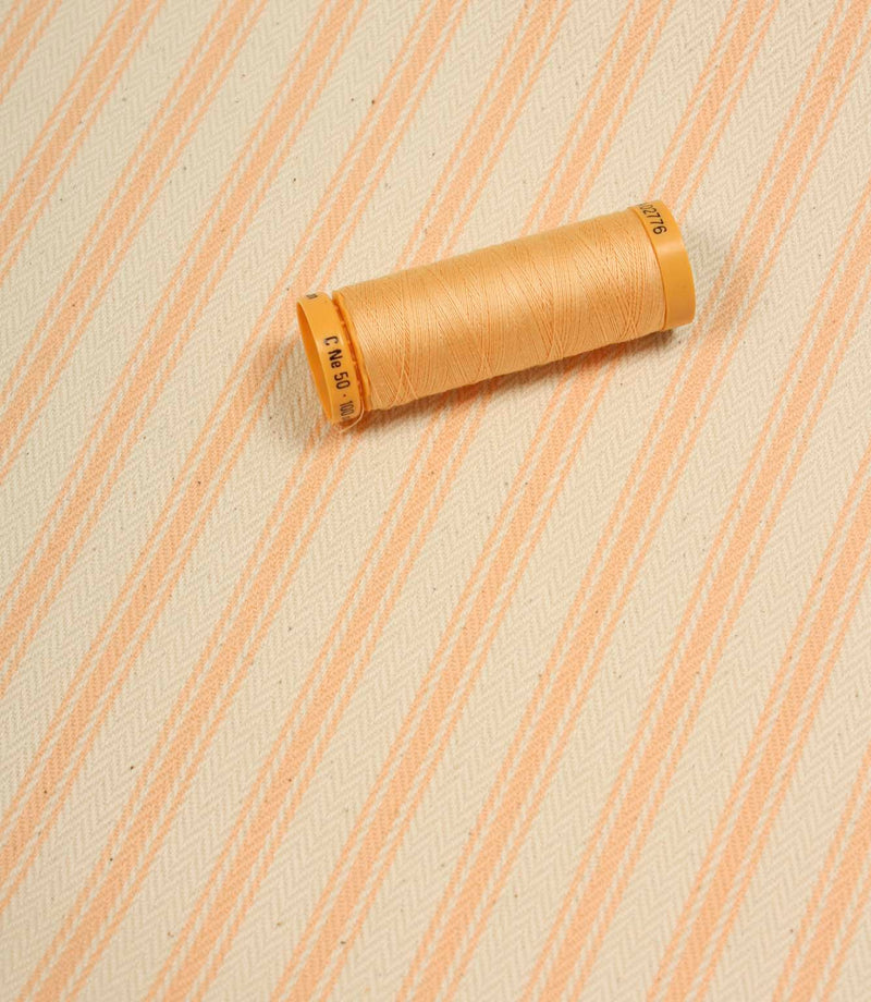 Peach ticking stripe fabric displayed on the diagonal with matching peach thread spool for colour reference.
