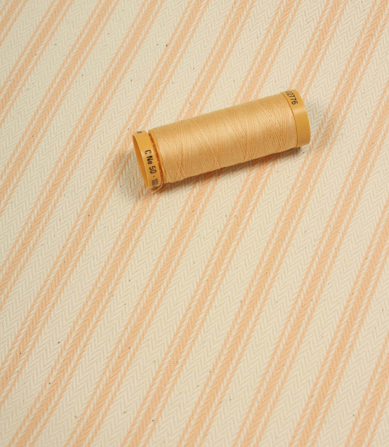 Peach ticking stripe fabric displayed on the diagonal with matching peach thread spool for colour reference.