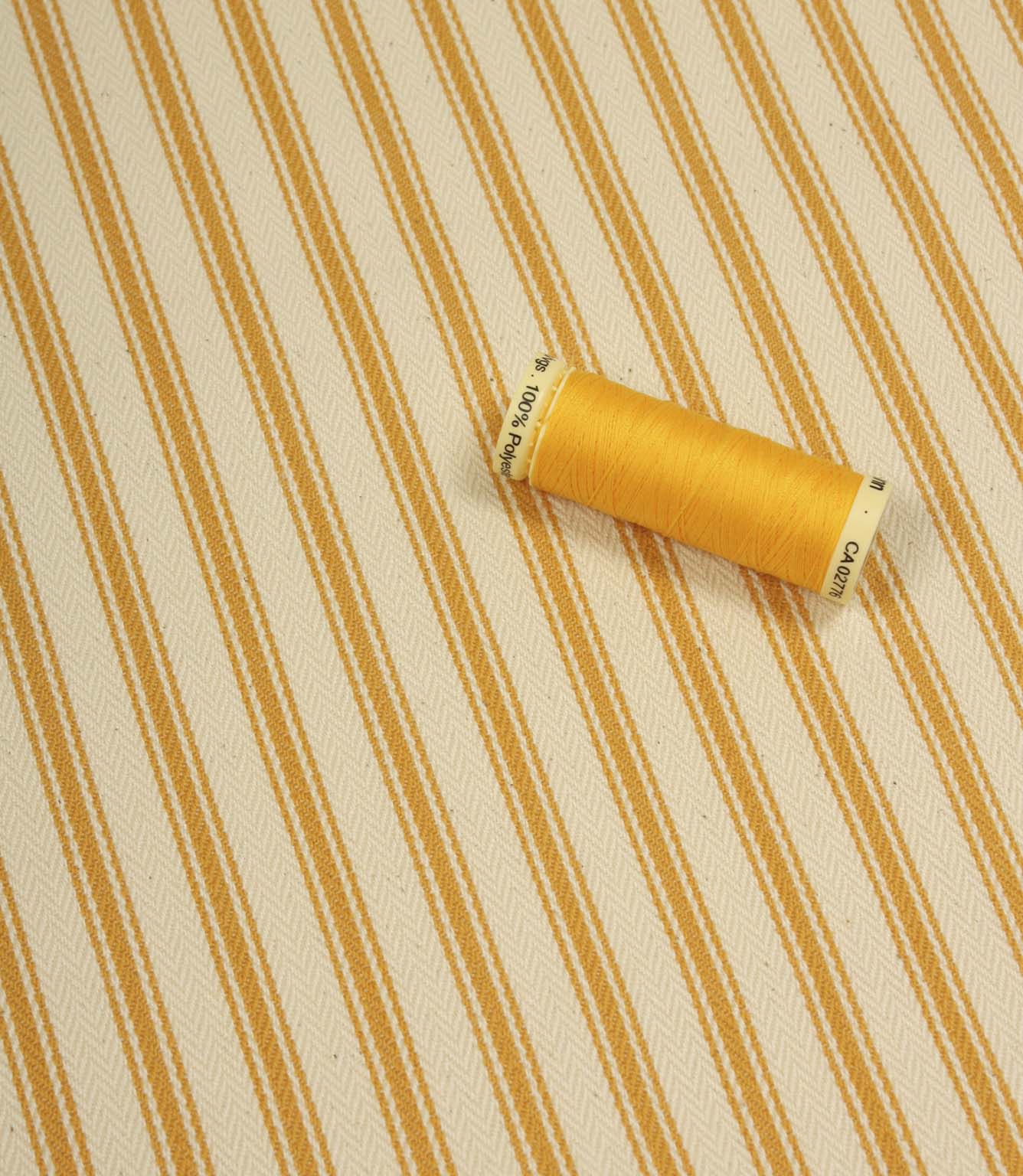 Turmeric yellow ticking stripe fabric shown on the diagonal with matching yellow thread spool for colour reference.