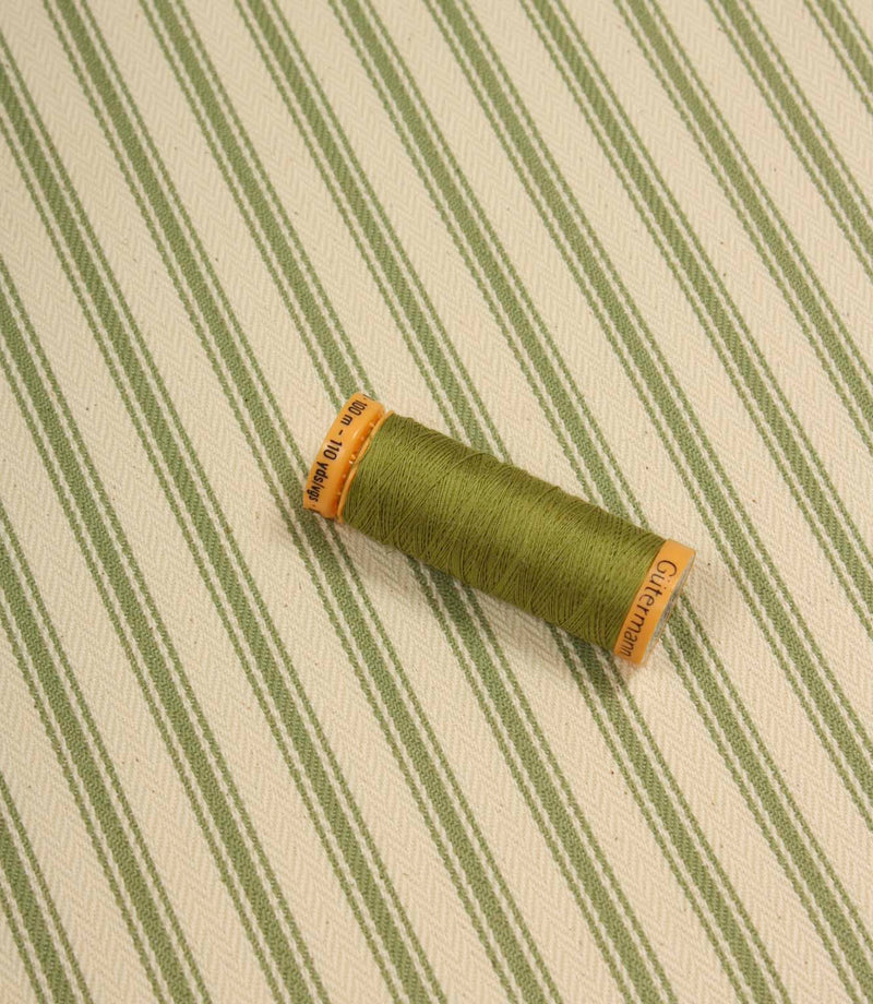 Warm green ticking stripe fabric displayed on the diagonal with matching green thread spool for colour reference.