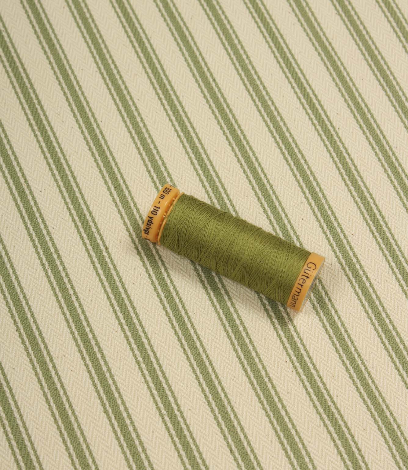Warm green ticking stripe fabric displayed on the diagonal with matching green thread spool for colour reference.