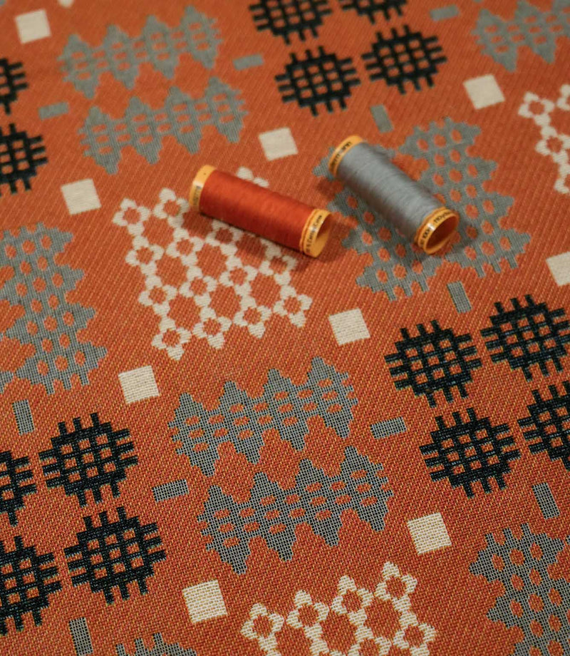 Kinfolk geometric fabric laid flat with orange and grey thread spools resting on the surface, highlighting the fabric’s woven texture and pattern.