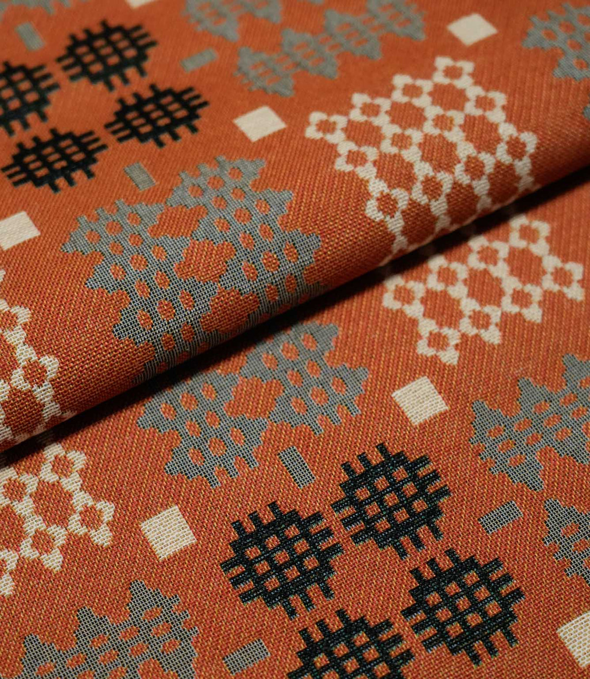 Folded Kinfolk fabric showing repeating Scandi-inspired geometric motifs in grey, black and cream against a warm terracotta orange ground.