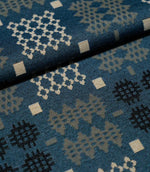 A close-up of patterned fabric with a blue base and geometric motifs in cream, light gray, and black, folded diagonally across the frame.