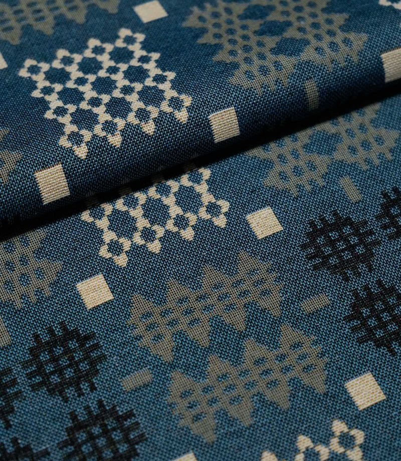 A close-up of patterned fabric with a blue base and geometric motifs in cream, light gray, and black, folded diagonally across the frame.