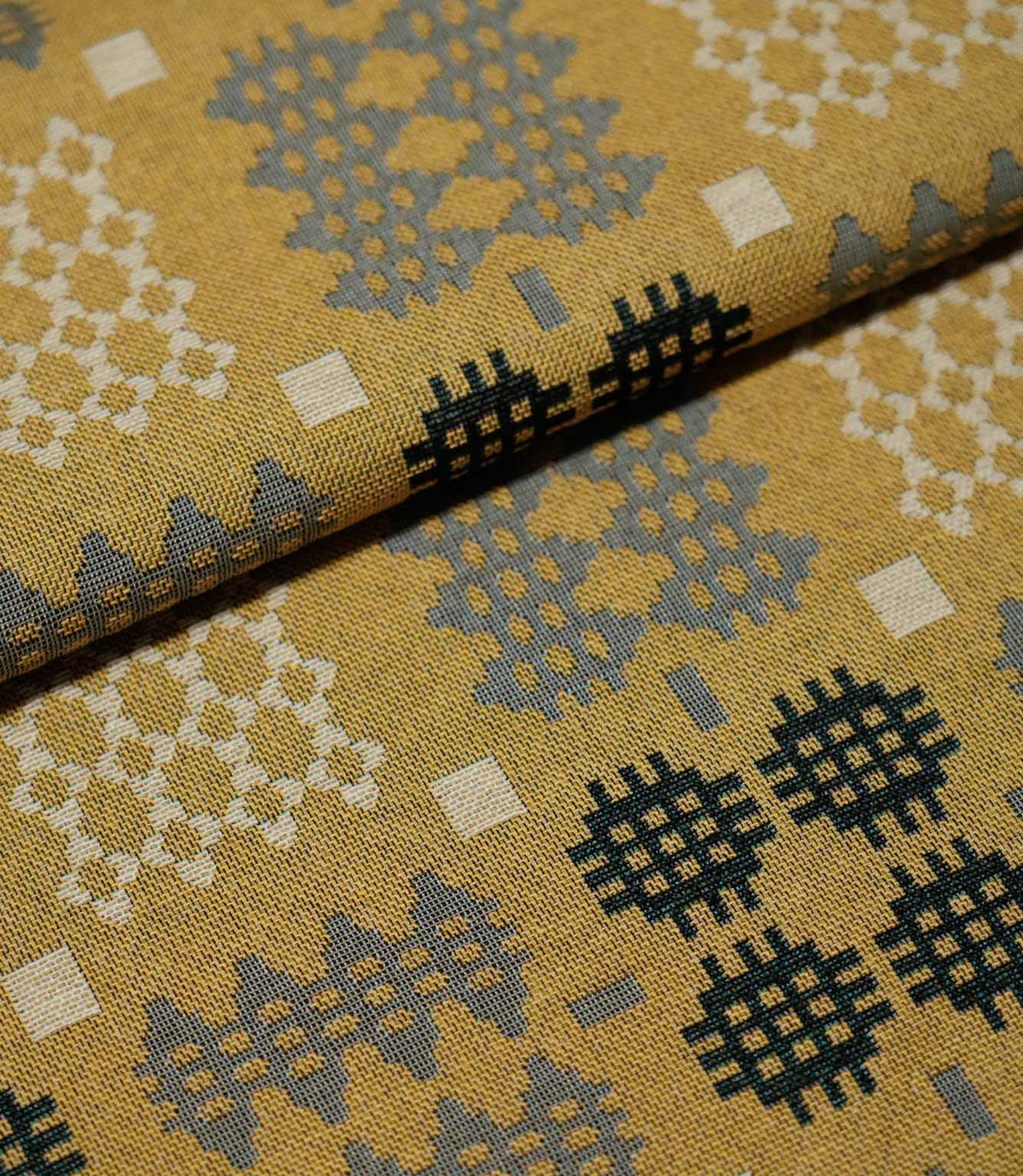 Folded Kinfolk fabric showing the scale and detail of its geometric pattern, with charcoal, grey and cream motifs on a warm mustard yellow background.
